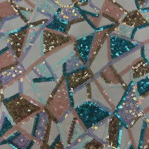 May include: A close-up of a fabric with a geometric pattern made of sequins. The sequins are in shades of blue, pink, and gold. The fabric is on a white background.