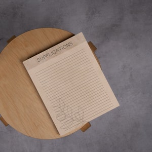 May include: A lined notebook with the word "SUPPLICATIONS" printed on the cover. The notebook has a simple floral design in the bottom right corner.