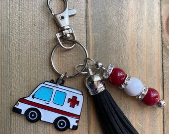 May include: A silver keychain with a white and red ambulance charm, a black tassel, and a beaded accent with red and white beads. The keychain has a lobster clasp for easy attachment.