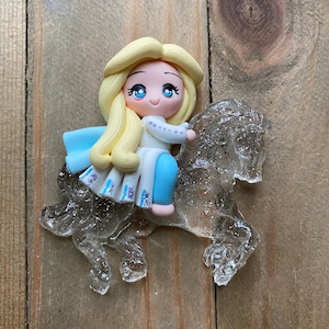 May include: A small, hand-crafted figurine of a blonde-haired girl riding a clear, translucent horse. The girl is wearing a white dress with blue accents and a blue cape. The figurine is made of polymer clay and is approximately 2 inches tall.
