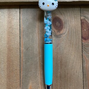 Op de afbeelding: Een blauwe pen met een Hello Kitty-figuur op de bovenkant. De pen heeft een doorzichtig gedeelte met glitter en kleine Hello Kitty-bedels erin.
