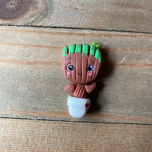 May include: A small, brown, clay figurine of a character from the Guardians of the Galaxy movie. The character is a tree-like humanoid with green leaves on its head and a white pot with a red heart on the side. The figurine has a smiling face with large eyes and a small mouth.