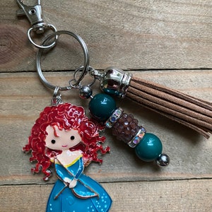 May include: A keychain featuring a cartoon character with red hair and a blue dress, holding a bow and arrow. The keychain also includes a brown tassel, green beads, and a silver charm.