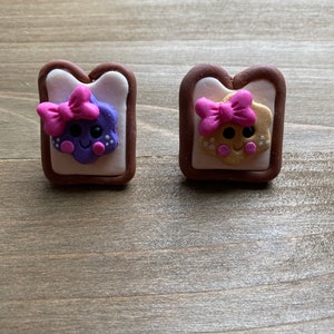 May include: Two small, handmade clay charms shaped like pieces of toast with cute faces. One charm has purple jam and the other has a brown peanut butter filling. Both charms have pink bows and white eyes.