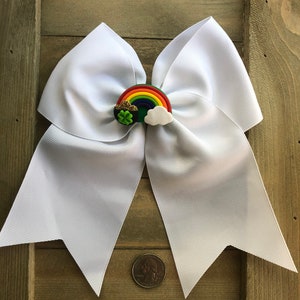 May include: White hair bow with a rainbow, shamrock, and cloud embellishment. The bow is approximately the size of a UK 20 pence coin.