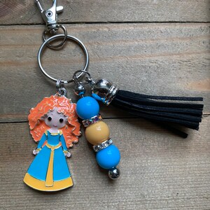 May include: A keychain featuring a cartoon character with red hair, a blue dress, and a yellow sash. The keychain also includes a black tassel and three beads: blue, yellow, and blue.