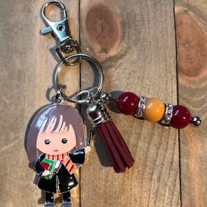 May include: A silver key chain with a charm of a cartoon girl wearing a black robe, a red and yellow scarf, and holding a book. The charm is attached to a silver ring with a burgundy tassel and a beaded charm with red, orange, and clear beads.