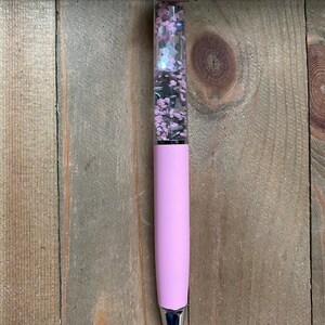 以下が含まれることがあります： ピンクのペンで、透明な部分にはピンクと銀色のグリッターが詰まっています。ペンの上部は、ピンクのカップケーキを着た灰色の猫の置物です。