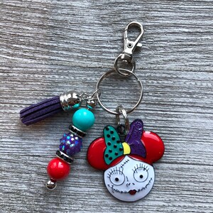 May include: A keychain featuring a cartoon character with red hair, a purple bow, and a white face with black eyes and a stitched mouth. The keychain also includes a purple tassel, a turquoise bead, and a red bead with silver accents.