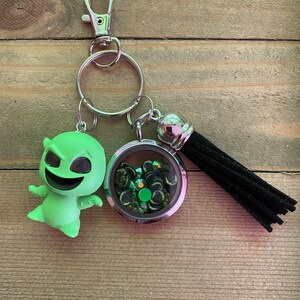 May include: Green character keychain with a silver charm containing green and black glitter. The charm is attached to a black tassel.