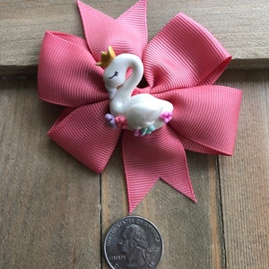 May include: A pink hair bow with a white swan figurine wearing a gold crown. The bow is made of grosgrain ribbon and has a quarter next to it for size comparison. The swan is decorated with small flowers.