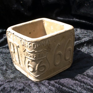 May include: A square concrete planter with the words "California Route 66" embossed on the sides. The planter is a light beige color.