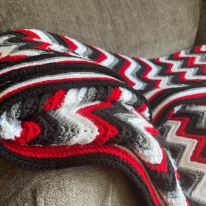 May include: A handmade crochet blanket with a bold zigzag pattern. The blanket features a striking color scheme of red, black, white, and gray. The blanket is folded over a brown couch, showcasing its texture and design.