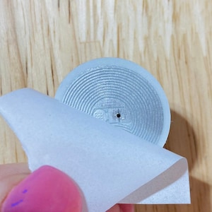 May include: A silver RFID tag with a circular shape and a small hole in the centre. The tag is being peeled off of a white backing.