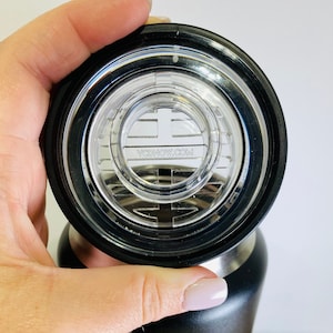 Può includere: Una borraccia isolante nera con coperchio trasparente. Il coperchio ha un bordo nero e il testo VCDNOW.COM. La bottiglia è cilindrica e ha una finitura opaca. Il coperchio è aperto.