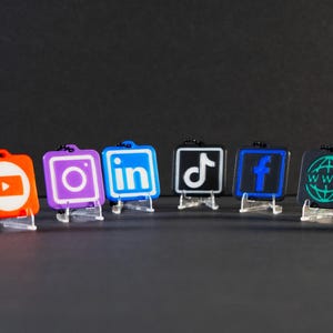 May include: A collection of small, square-shaped keychains featuring social media logos. The keychains are in various colors, including red, purple, blue, black, and green, with white and neon-colored icons. Each keychain is displayed on a clear plastic stand.