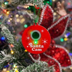 May include: A red, circular Christmas ornament with the text "Santa Cam" and snowflake designs. The ornament has a green center and a green light. It is hanging on a Christmas tree with red bows and lights.