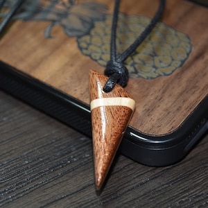 Puede incluir: Un colgante de madera con un patrón de veta de madera marrón claro y marrón oscuro. El colgante tiene forma de punta afilada y está unido a un cordón negro.