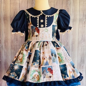 May include: A blue and white dress with a Christmas nativity scene print. The dress has a white lace collar and a blue lace trim. The dress is on a mannequin.