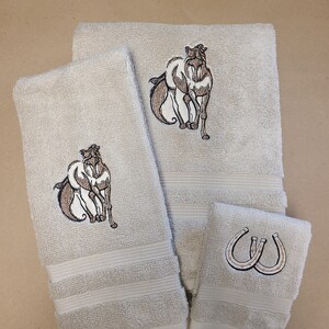 May include: A set of three beige towels with embroidered horse and horseshoe designs. The towels are made of soft, absorbent cotton and are perfect for everyday use.