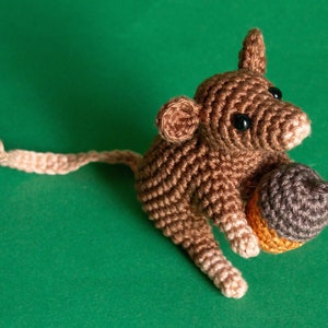 Puede incluir: Un ratón de crochet marrón sosteniendo una bellota marrón y beige. El ratón está sentado sobre una superficie verde.