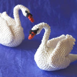Puede incluir: Dos cisnes blancos de ganchillo hechos a mano con picos negros y detalles rojos sobre un fondo azul. Los cisnes están colocados uno frente al otro, mostrando su detallada artesanía. Un encantador artículo decorativo.