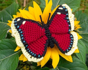 May include: A crocheted butterfly with red wings, black accents, and a yellow and white border, sitting on a yellow sunflower.