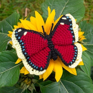 Puede incluir: Una mariposa de crochet con alas rojas, detalles negros y un borde amarillo y blanco, sentada sobre un girasol amarillo.