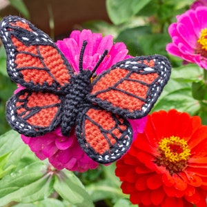 May include: A crocheted butterfly with black, red, and white accents is perched on a pink zinnia flower. The butterfly is a popular craft project and a great addition to any home decor.