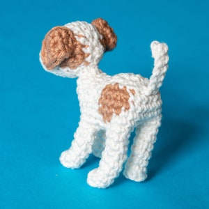 May include: A crocheted white dog with brown spots. The dog is standing on a blue background.