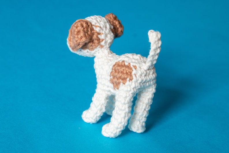 May include: A crocheted white dog with brown spots. The dog is standing on a blue background.