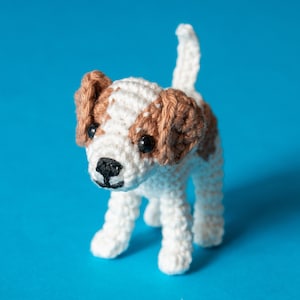 May include: A crocheted white and brown dog toy with black eyes. The dog is standing on a blue background.