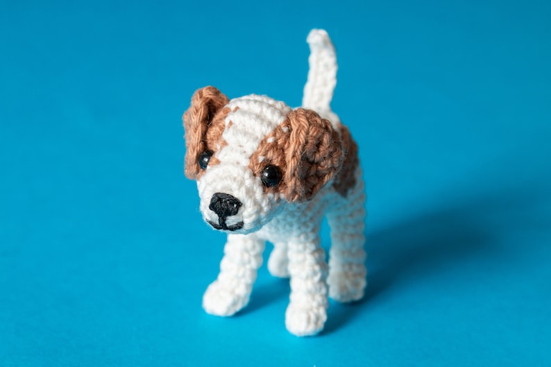 May include: A crocheted white and brown dog toy with black eyes. The dog is standing on a blue background.