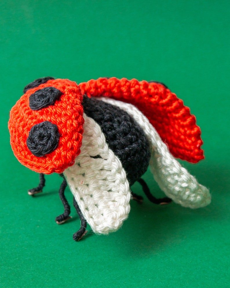Puede incluir: Una mariquita de ganchillo hecha a mano con cabeza roja con lunares negros, cuerpo negro y alas blancas y rojas. La mariquita tiene patas negras, sobre un fondo verde.