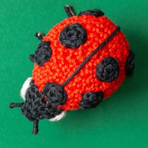 Puede incluir: Una mariquita de ganchillo hecha a mano, roja con lunares negros y cabeza negra. La mariquita tiene peque&ntilde;as antenas y est&aacute; sobre un fondo verde. Ideal para juguetes infantiles o decoraci&oacute;n del hogar.