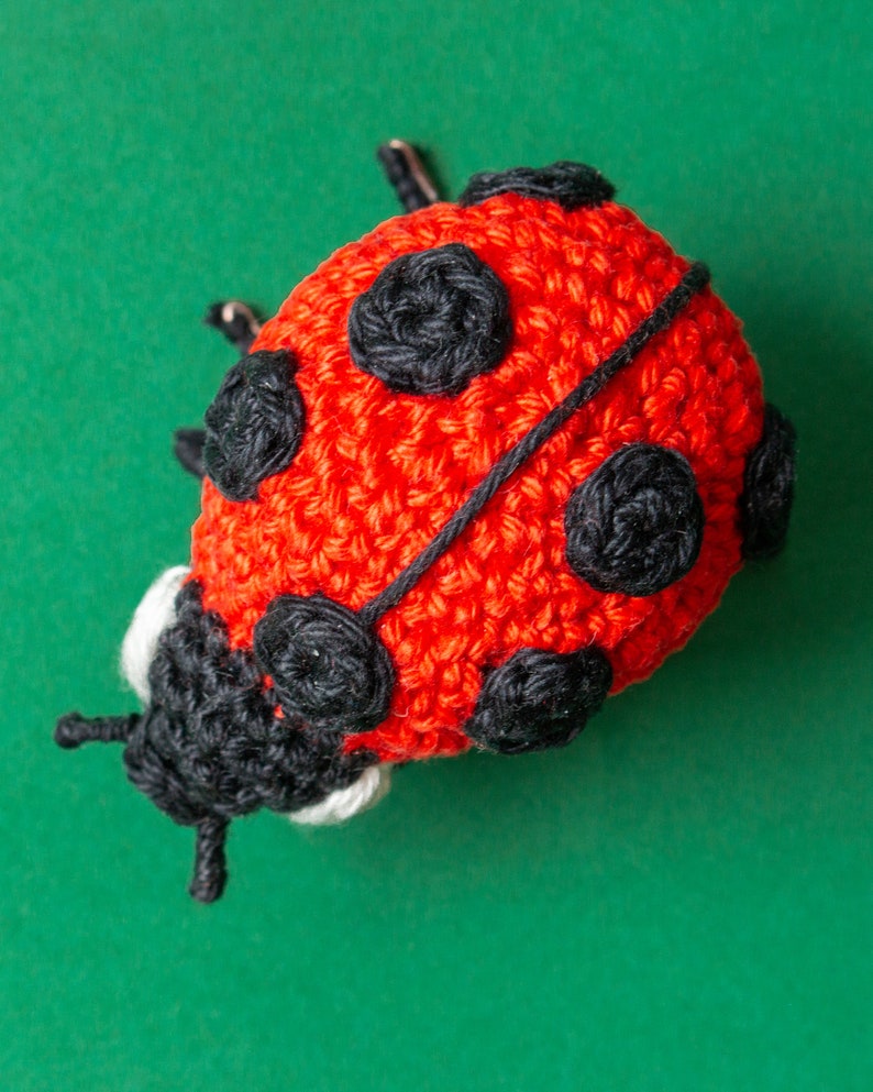 Puede incluir: Una mariquita de ganchillo hecha a mano, roja con lunares negros y cabeza negra. La mariquita tiene peque&ntilde;as antenas y est&aacute; sobre un fondo verde. Ideal para juguetes infantiles o decoraci&oacute;n del hogar.