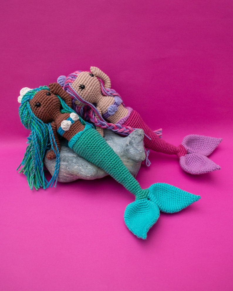 May include: Two crocheted mermaid dolls with long hair and tails. One mermaid is brown with teal hair and a turquoise tail. The other mermaid is light brown with pink hair and a purple tail. The mermaids are sitting on a gray rock against a pink background.
