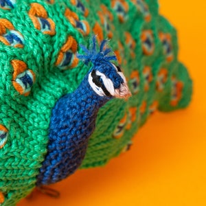 K&ouml;nnte beinhalten: Ein handgestrickter Pfau mit einem leuchtend gr&uuml;nen Schwanz und einem blauen Kopf. Der Schwanz hat orange und blaue Akzente, vor einem hellorangen Hintergrund. Dieser dekorative Artikel ist wahrscheinlich ein Stofftier oder ein Einrichtungsgegenstand.