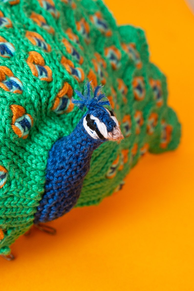 K&ouml;nnte beinhalten: Ein handgestrickter Pfau mit einem leuchtend gr&uuml;nen Schwanz und einem blauen Kopf. Der Schwanz hat orange und blaue Akzente, vor einem hellorangen Hintergrund. Dieser dekorative Artikel ist wahrscheinlich ein Stofftier oder ein Einrichtungsgegenstand.