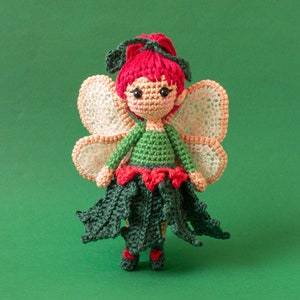 May include: A crocheted fairy doll with red hair, green and red clothing, and white wings. The doll is standing on a green background.