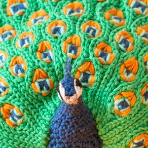 K&ouml;nnte beinhalten: Nahaufnahme eines gestrickten Pfauen-Designs. Der Pfau hat einen blauen K&ouml;rper und Kopf mit einem wei&szlig;en und schwarzen Gesicht. Die Schwanzfedern sind gr&uuml;n mit orangefarbenen und blauen Akzenten. Die Strickstruktur ist im gesamten Design sichtbar.
