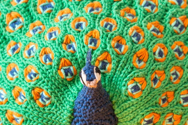 K&ouml;nnte beinhalten: Nahaufnahme eines gestrickten Pfauen-Designs. Der Pfau hat einen blauen K&ouml;rper und Kopf mit einem wei&szlig;en und schwarzen Gesicht. Die Schwanzfedern sind gr&uuml;n mit orangefarbenen und blauen Akzenten. Die Strickstruktur ist im gesamten Design sichtbar.