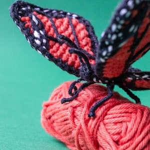 May include: A crocheted butterfly with black and red wings perched on a ball of red yarn.