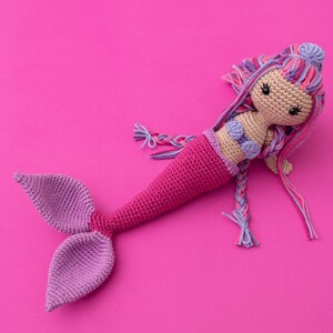 May include: A crocheted mermaid doll with a pink tail, purple and pink hair, and a purple seashell bra. The doll is lying on a pink background.
