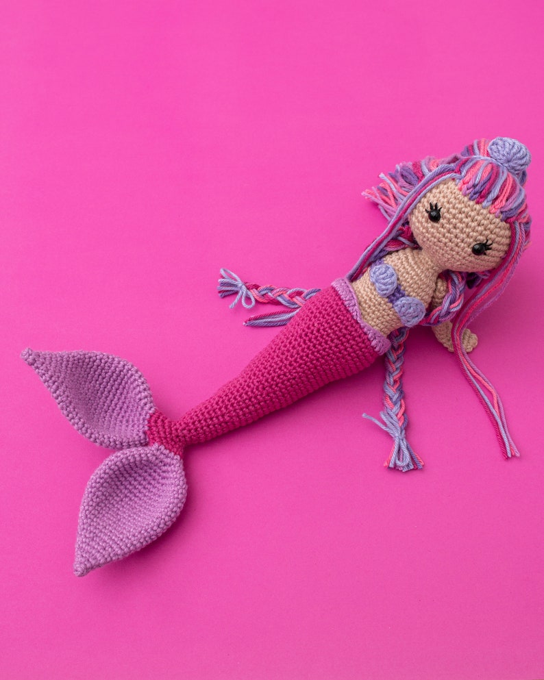 May include: A crocheted mermaid doll with a pink tail, purple and pink hair, and a purple seashell bra. The doll is lying on a pink background.