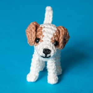 May include: A crocheted white and brown dog toy with black eyes and a black nose. The toy is standing on a blue background.