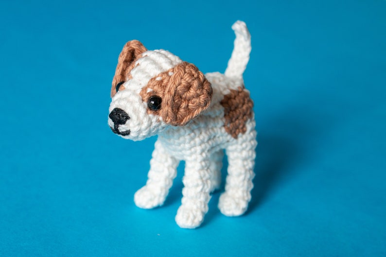 May include: A crocheted white and brown dog toy with black eyes. The dog is standing on a blue background.