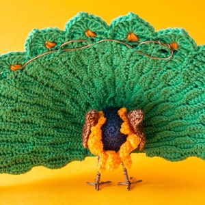 K&ouml;nnte beinhalten: Handgefertigte Pfauenfigur mit einem leuchtend gr&uuml;nen geh&auml;kelten Schwanz, akzentuiert mit orangefarbenen Details und Kupferdraht. Der K&ouml;rper hat blaue, gelbe und braune H&auml;kelarbeiten mit Drahtbeinen. Vor einem leuchtend gelben Hintergrund.