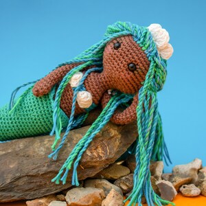 May include: A crocheted mermaid doll with brown skin, green and blue hair, and white seashells. The doll is sitting on a pile of rocks.