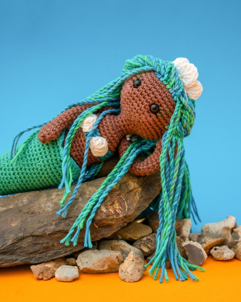 May include: A crocheted mermaid doll with brown skin, green and blue hair, and white seashells. The doll is sitting on a pile of rocks.